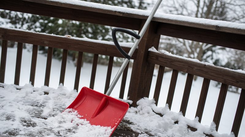 A red snow shovel with a second curved handle leaning on a snowy deck railing, in Flagstaff, Arizona